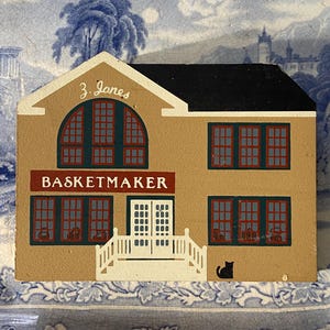 May include: A tan-coloured, folk art style building with a black roof and the text "BASKETMAKER" above the double doors. The building has red and green window frames and a small black cat silhouette on the side.