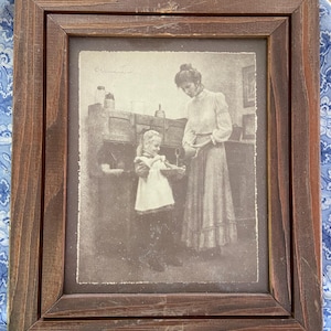 May include: A framed sepia-toned print depicting a woman and a young girl in a domestic setting. The print is encased in a dark brown wooden frame. The image shows the woman pouring from a container while the girl holds a garment. The frame is resting on a blue and white floral patterned cloth.