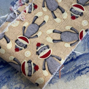 May include: Close-up of a fabric swatch featuring a repeating pattern of sock monkey illustrations. The monkeys are gray with red and white accents, set against a tan background with small, off-white circles. The fabric has a frayed edge.