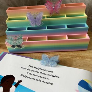May include: A tiered, multi-compartment organizer featuring a pastel rainbow gradient finish. Attached to the front are several translucent butterfly-shaped clips in pink, purple, teal, and blue. A book page with text is visible in the foreground.