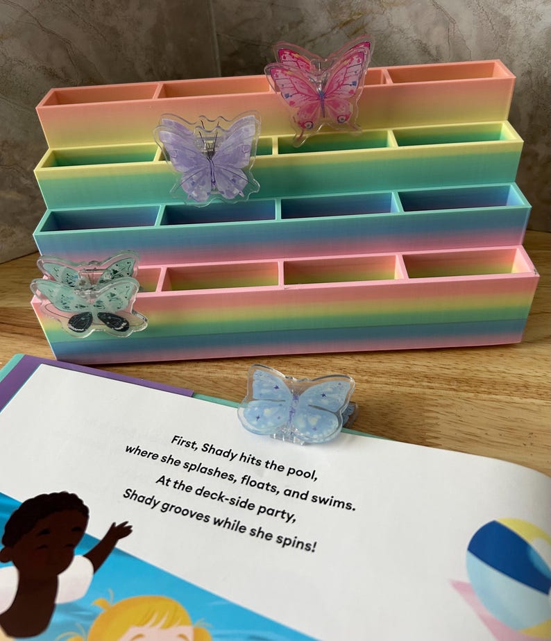 May include: A tiered, multi-compartment organizer featuring a pastel rainbow gradient finish. Attached to the front are several translucent butterfly-shaped clips in pink, purple, teal, and blue. A book page with text is visible in the foreground.