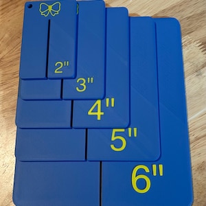 May include: A set of five blue rectangular plastic templates with rounded corners, each featuring a yellow printed number indicating sizes 2", 3", 4", 5", and 6". The smallest template includes a small circular hole and a yellow bow icon.