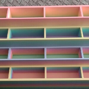 May include: A tiered display stand featuring four levels, each divided into four rectangular compartments. The structure exhibits a smooth gradient color transition, shifting through shades of pink, yellow, blue, and green across its surface.