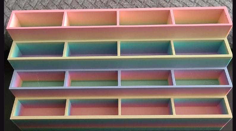 May include: A tiered display stand featuring four levels, each divided into four rectangular compartments. The structure exhibits a smooth gradient color transition, shifting through shades of pink, yellow, blue, and green across its surface.