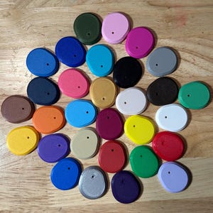 May include: A collection of small, irregularly shaped, flat plastic discs in a wide variety of solid colors including red, blue, green, yellow, pink, purple, orange, white, black, gray, and brown, each featuring a single small hole near the center.