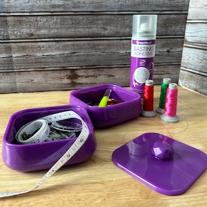 May include: Two purple square containers with lids, one containing sewing supplies like thread, needles, and a measuring tape, the other containing a yellow tool. A can of basting adhesive spray and three spools of thread are on a wooden surface.