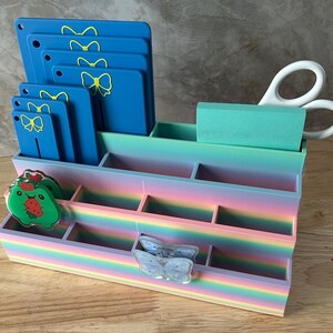 May include: A multi-tiered desk organizer with a rainbow gradient finish featuring several compartments. It holds blue rectangular tags with yellow bow icons, a green notepad, white scissors, a green strawberry character charm, and a butterfly clip.