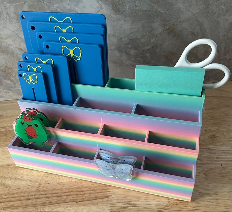 May include: A multi-tiered desk organizer with a rainbow gradient finish featuring several compartments. It holds blue rectangular tags with yellow bow icons, a green notepad, white scissors, a green strawberry character charm, and a butterfly clip.