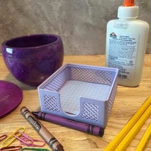 May include: A collection of school and office supplies on a wooden surface. Includes a purple bowl, a light purple desk organizer, a bottle of glue, crayons, paper clips, and pencils. The Crayola crayon is brown with the text "Crayola.com".