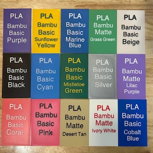 May include: A grid of fifteen rectangular PLA filament sample swatches in various colors including purple, yellow, blue, green, beige, black, cyan, silver, lilac, coral, pink, tan, white, and cobalt, each labeled with its specific color name.