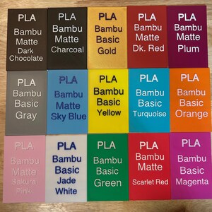 May include: A collection of fifteen rectangular PLA filament sample swatches arranged in a grid. Each swatch features embossed text identifying the brand, material type, and specific color name, including shades like Dark Chocolate, Charcoal, Gold, and Magenta.
