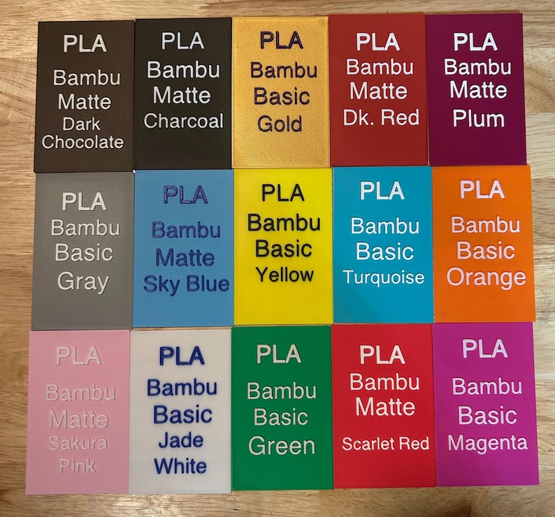 May include: A collection of fifteen rectangular PLA filament sample swatches arranged in a grid. Each swatch features embossed text identifying the brand, material type, and specific color name, including shades like Dark Chocolate, Charcoal, Gold, and Magenta.