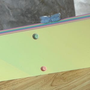 May include: A rectangular clutch bag with a pale yellow base and a multi-colored pastel striped top edge. The bag features a light blue butterfly-shaped clasp on the top and five small circular feet on the bottom surface for stability.