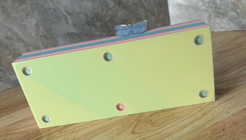 May include: A rectangular clutch bag with a pale yellow base and a multi-colored pastel striped top edge. The bag features a light blue butterfly-shaped clasp on the top and five small circular feet on the bottom surface for stability.