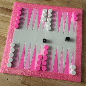 May include: A pink and white backgammon board with pink glitter edging. The board features pink and white triangles, with pink and white game pieces, and two black dice. A fun game for family and friends.