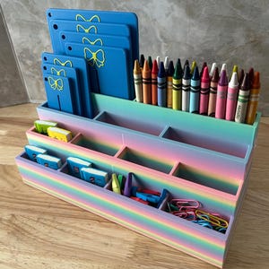 May include: A tiered desk organizer with a rainbow gradient finish containing various compartments. It holds a row of colorful crayons, several blue rectangular boards with yellow bow icons, small miniature books, and assorted metal paper clips.