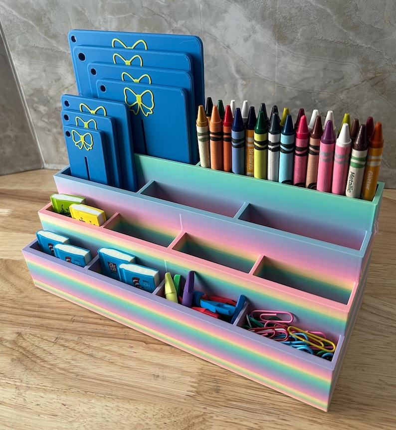 May include: A tiered desk organizer with a rainbow gradient finish containing various compartments. It holds a row of colorful crayons, several blue rectangular boards with yellow bow icons, small miniature books, and assorted metal paper clips.