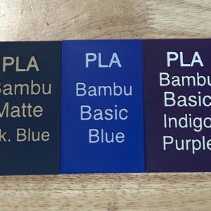 May include: Five rectangular PLA filament swatches in brown, dark blue, blue, indigo purple, and red. Each swatch features white printed text identifying the material type, brand, and specific color name, such as PLA Bambu Basic Brown and PLA Bambu Matte Dk. Blue.