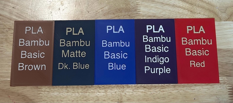 May include: Five rectangular PLA filament swatches in brown, dark blue, blue, indigo purple, and red. Each swatch features white printed text identifying the material type, brand, and specific color name, such as PLA Bambu Basic Brown and PLA Bambu Matte Dk. Blue.