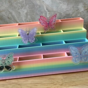 May include: A tiered desk organizer featuring a pastel rainbow gradient finish. The unit has four levels with multiple rectangular compartments. Four translucent butterfly-shaped accents in pink, purple, and blue are attached to the front panels.