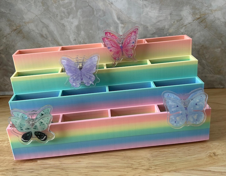 May include: A tiered desk organizer featuring a pastel rainbow gradient finish. The unit has four levels with multiple rectangular compartments. Four translucent butterfly-shaped accents in pink, purple, and blue are attached to the front panels.