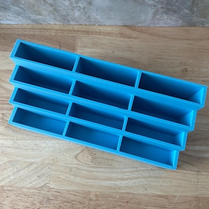May include: A bright blue tiered organizer featuring four horizontal rows, each divided into three rectangular compartments. The structure is stepped, with each row positioned slightly lower than the one behind it, creating a cascading display shelf.