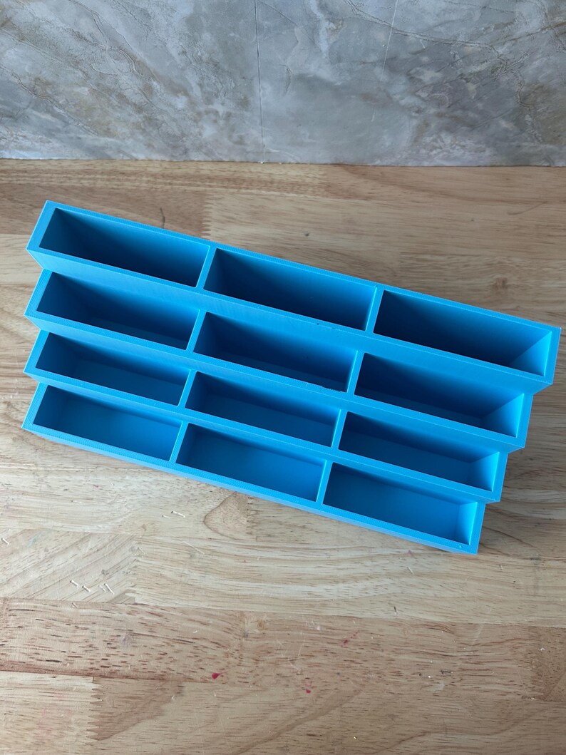 May include: A bright blue tiered organizer featuring four horizontal rows, each divided into three rectangular compartments. The structure is stepped, with each row positioned slightly lower than the one behind it, creating a cascading display shelf.