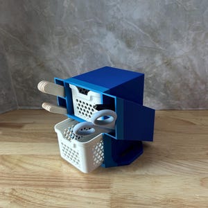 May include: A blue 3D-printed desk organizer featuring two stacked white plastic baskets. The top basket holds wooden craft sticks, while the bottom basket contains a pair of light gray scissors and a black pen. The unit has a solid blue frame.