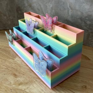 May include: A tiered, rainbow-colored desk organizer with multiple rectangular compartments. The organizer features several translucent, butterfly-shaped decorative accents in shades of pink, purple, and blue attached to the front edges.