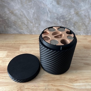 May include: A black coin organizer with a spiral design and a removable lid. The top of the organizer has several coin slots, some filled with coins. The organizer is on a wooden surface.