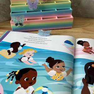 May include: A tiered, rainbow-colored desk organizer with three butterfly-shaped clips attached to the front. Behind it lies an open children's book featuring a colorful illustration of diverse characters swimming in a pool with text about Shady.