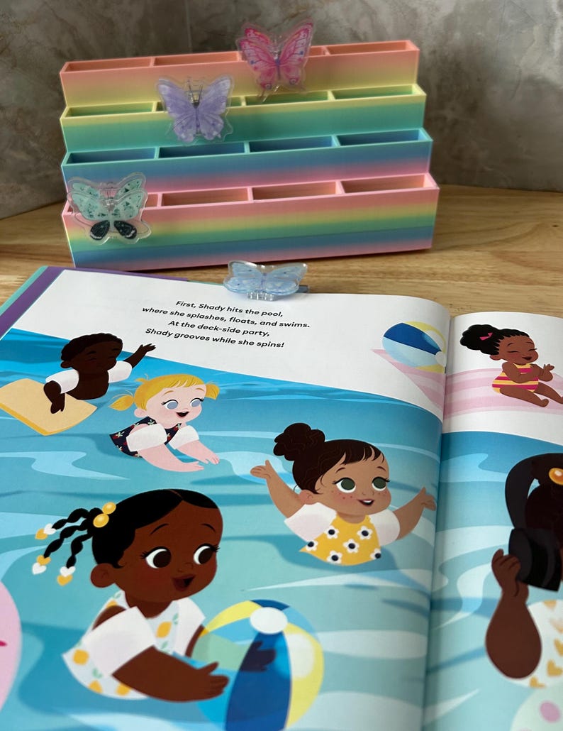 May include: A tiered, rainbow-colored desk organizer with three butterfly-shaped clips attached to the front. Behind it lies an open children's book featuring a colorful illustration of diverse characters swimming in a pool with text about Shady.