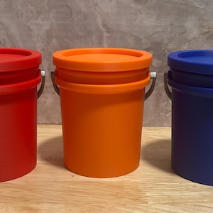 May include: Three cylindrical plastic buckets with lids and metal handles, arranged in a row. The buckets are solid red, orange, and blue. Each bucket features a smooth matte finish, a circular lid, and a sturdy metal bail handle attached to the sides.