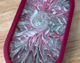 May include: An oval-shaped tray featuring a deep magenta border and a central resin inlay. The inlay displays a swirling, marbled pattern of soft pink, white, and pale green hues that radiate outward, creating a complex, floral-like texture.