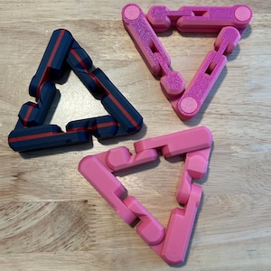 May include: Three geometric triangle puzzles in shades of pink and navy blue. One puzzle is navy blue with red stripes, another is a glittery pink, and the third is a solid pink color. The puzzles are on a wooden surface.