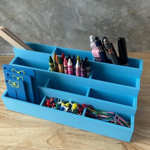 May include: A tiered light blue desk organizer with multiple compartments containing wooden craft sticks, assorted crayons, pens, push pins, paper clips, and three blue rectangular tags featuring yellow bow icons on the front left side.