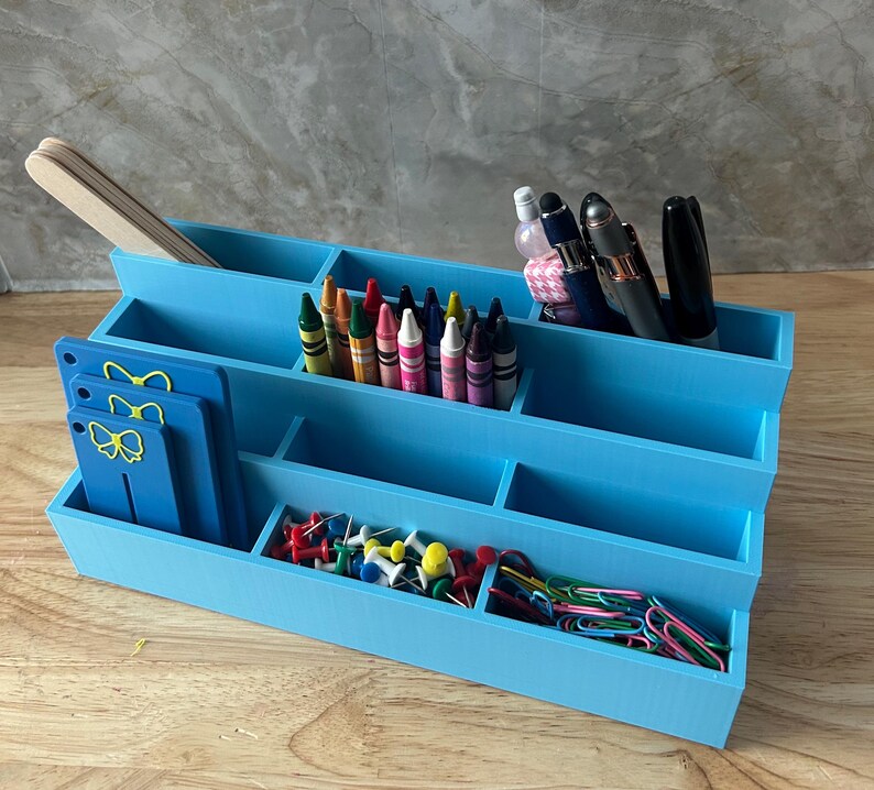 May include: A tiered light blue desk organizer with multiple compartments containing wooden craft sticks, assorted crayons, pens, push pins, paper clips, and three blue rectangular tags featuring yellow bow icons on the front left side.