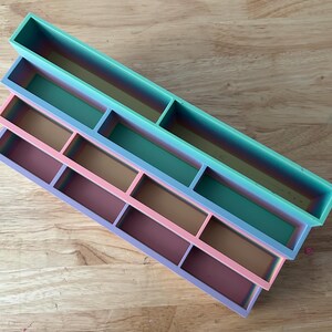 May include: A set of three rectangular, tiered storage trays featuring a gradient color scheme. The top tray is teal, the middle tray transitions from teal to pink, and the bottom tray is a blend of pink and purple, each with internal dividers.