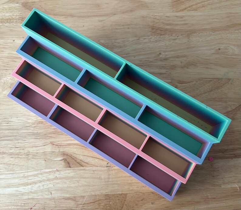 May include: A set of three rectangular, tiered storage trays featuring a gradient color scheme. The top tray is teal, the middle tray transitions from teal to pink, and the bottom tray is a blend of pink and purple, each with internal dividers.