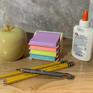 May include: A yellow apple-shaped container, a stack of purple, orange, lime green, and teal sticky notes in a holder, a white bottle of Elmer's school glue with an orange cap, two yellow pencils, and a gray ballpoint pen with rose gold accents.