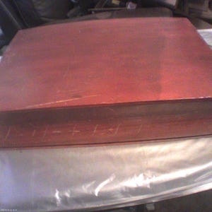 May include: A rectangular wooden box with a smooth, reddish-brown finish. The lid is flat and covers the entire top surface. The front side of the box features a series of faint, horizontal, linear scratch marks across the wood grain.