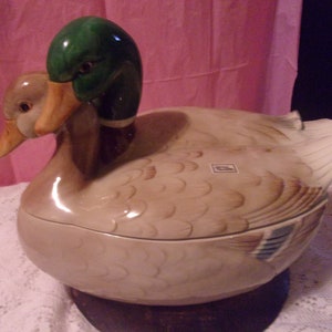May include: A ceramic duck-shaped serving dish with a lid, featuring two duck heads. The duck's head is green and brown, with a tan body and wings. The dish is likely used for serving food and adds a decorative touch to the table.