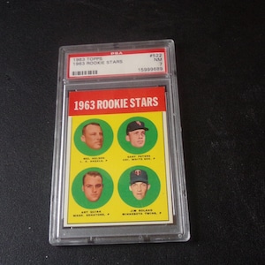 Peut inclure: Une carte de baseball 1963 Topps Rookie Stars, dans un étui de protection. La carte présente quatre portraits de joueurs dans des cercles verts sur un fond jaune, avec le texte "1963 ROOKIE STARS" en haut.