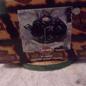 Pode incluir: Uma chaleira de couro Harley Davidson preta com o logotipo da Harley Davidson na frente. A chaleira está sentada sobre uma base circular verde e marrom. A caixa em que a chaleira veio está no fundo e tem o logotipo da Harley Davidson.