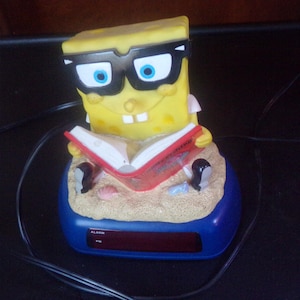 Può includere: Una sveglia gialla di SpongeBob SquarePants con occhiali neri, che legge un libro rosso. L'orologio è posizionato su una base blu con un display digitale. L'orologio ha un design a spiaggia sabbiosa.