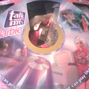 Może przedstawiać: Płyta CD-ROM Talk With Me Barbie w oryginalnym okrągłym opakowaniu. Na płycie znajdują się grafiki przedstawiające blond lalkę, komputer oraz młodą osobę. Środek opakowania posiada okrągłe wycięcie, przez które widać lalkę wewnątrz.
