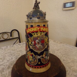 May include: A ceramic beer stein with a blue, yellow, and red design featuring the Looney Tunes characters. The stein has a pewter lid with a crown and a figure of Bugs Bunny in a knight's costume.
