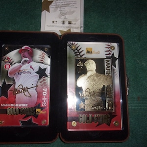 May include: A black and brown display case with two baseball cards inside. The card on the left features a photo of Mark McGwire in a St. Louis Cardinals uniform with the text "Mark McGwire Gold Stars 70 HRS". The card on the right features a gold-toned photo of Mark McGwire with the text "Mark McGwire Gold Stars".