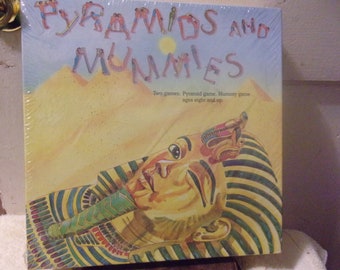 Könnte beinhalten: Eine Vintage-Spieleschachtel mit einer gelben, orangen und grünen Illustration eines Pharaos, der auf einem gestreiften Bett liegt. Die Schachtel trägt den Titel "PYRAMIDS AND MUMMIES" in großen, bunten Buchstaben. Der Text "Two games: Pyramid game, Mummy game. ages eight and up." ist unter dem Titel gedruckt.