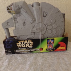 Op de afbeelding: Een grijze plastic Millennium Falcon draagkoffer met een Star Wars: The Power of the Force logo. De koffer is open en laat het interieur zien, dat is ontworpen om actionfiguren in te bewaren. De koffer bevat ook een exclusieve Han Solo actionfiguur.